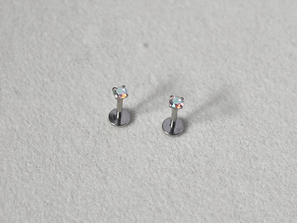 A pair of silver-toned labret earrings with small heart-shaped clear gemstones, displayed on a textured light gray surface.