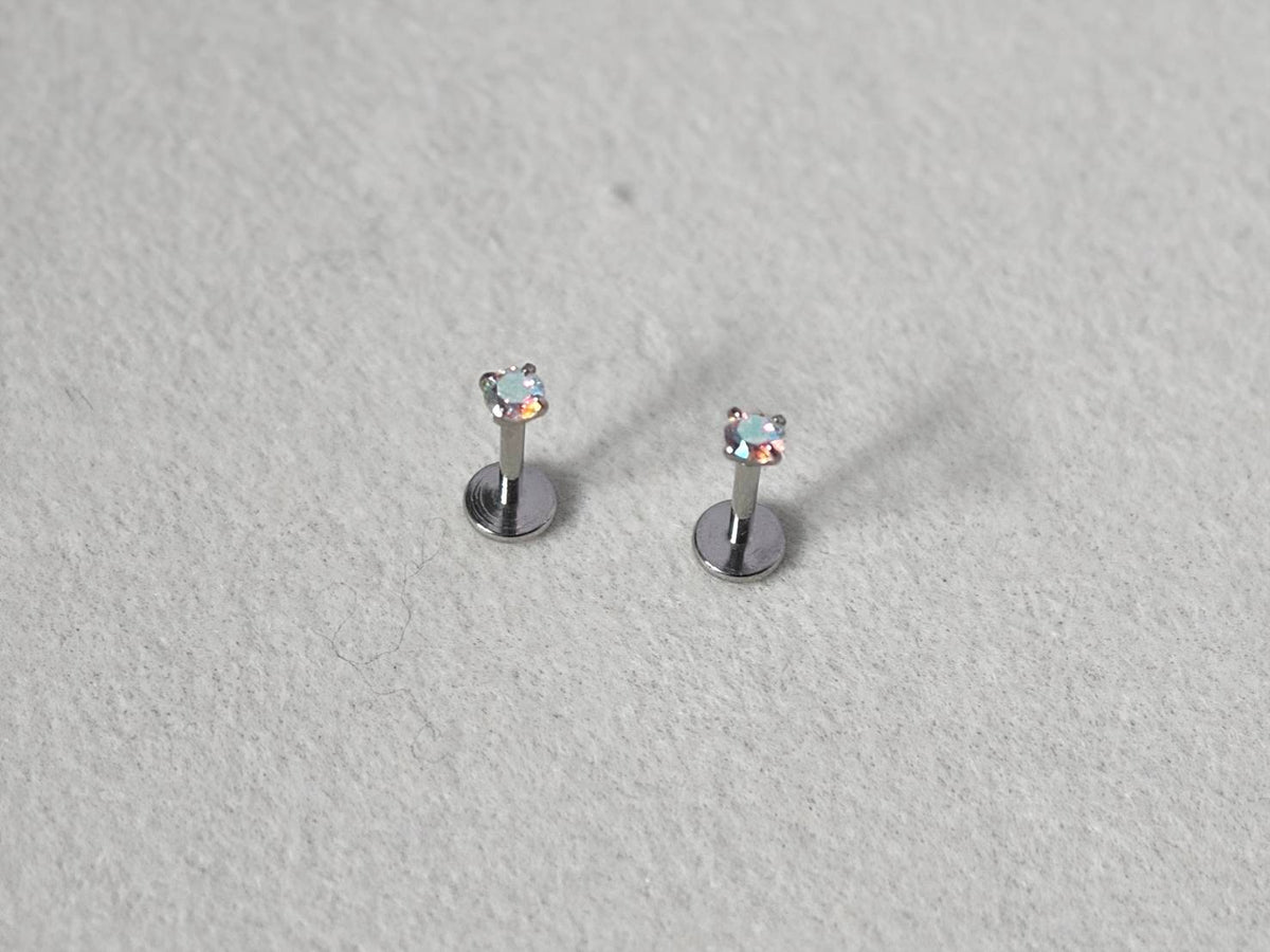 A pair of silver-toned labret earrings with small heart-shaped clear gemstones, displayed on a textured light gray surface.