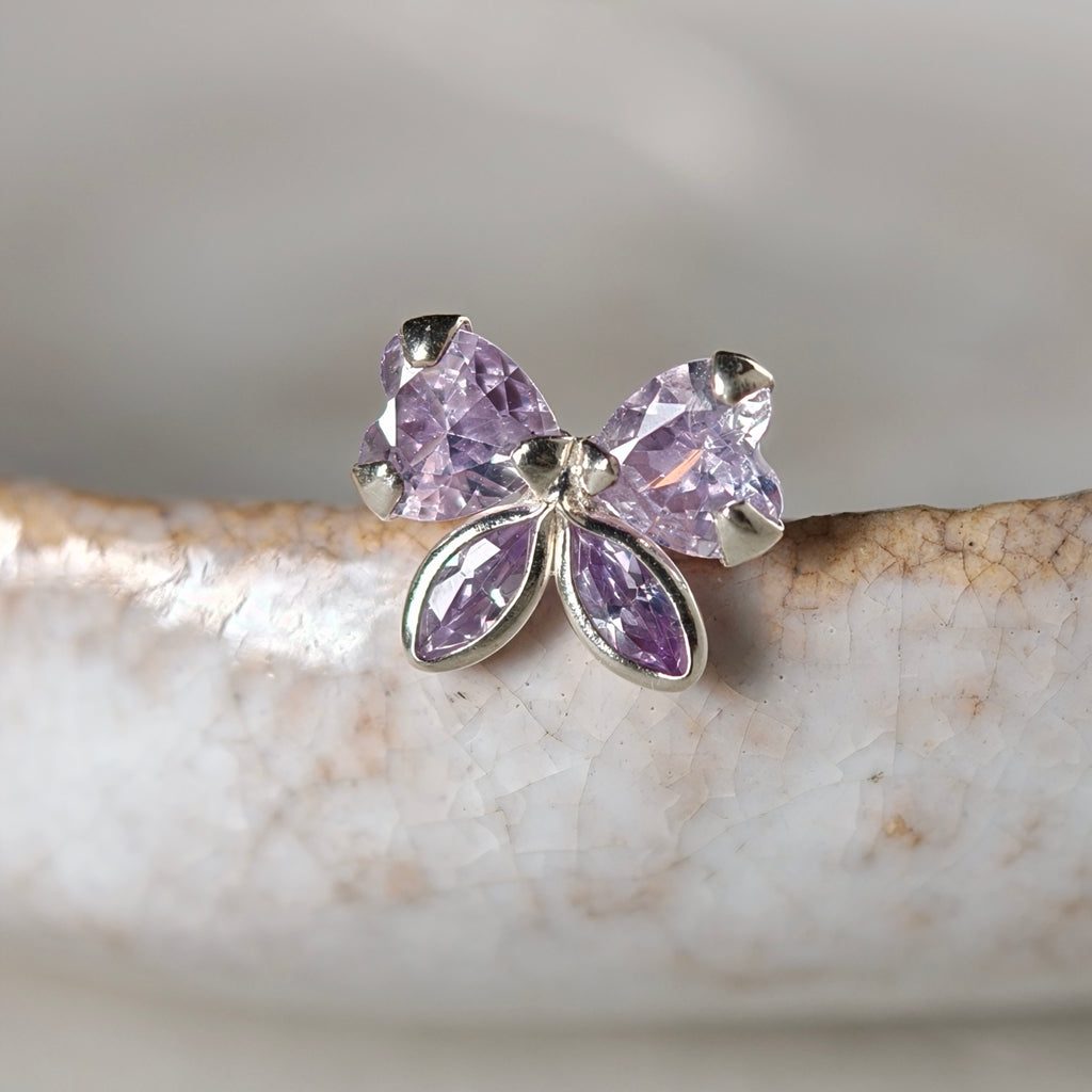 A silver butterfly stud earring with four faceted light purple gemstones, resting on a textured white ceramic surface.