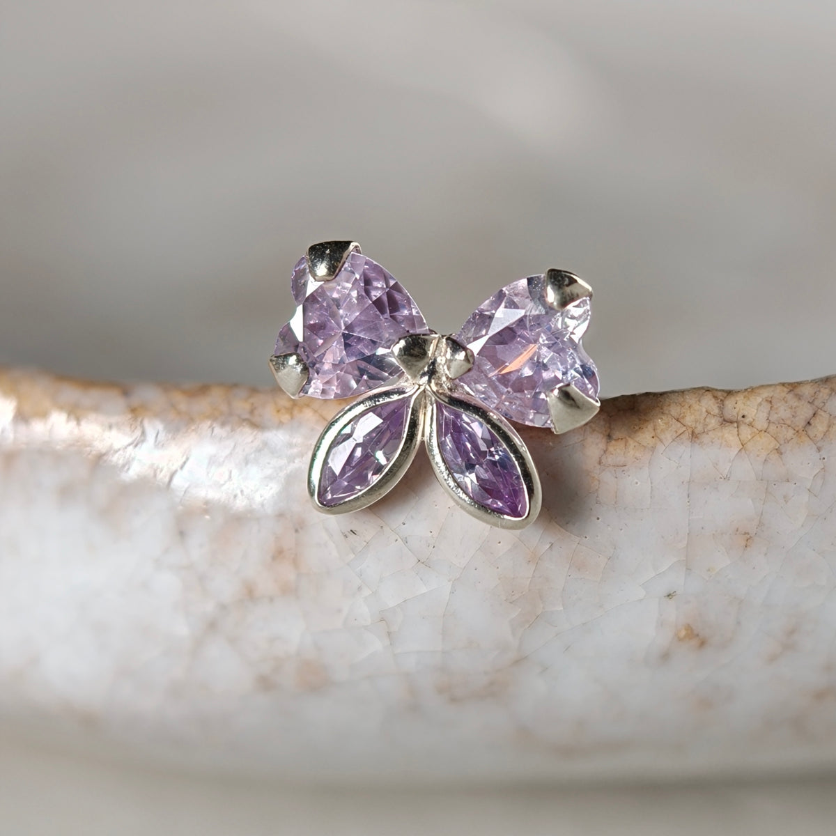 A silver butterfly stud earring with four faceted light purple gemstones, resting on a textured white ceramic surface.
